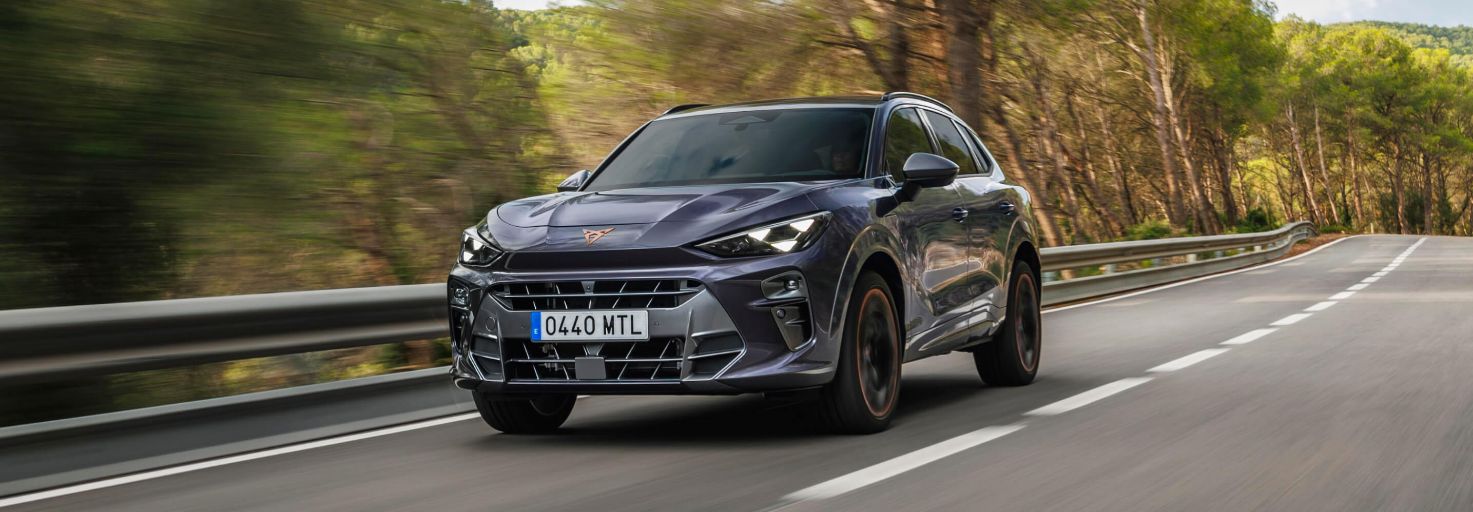 A dark grey Cupra Terramar SUV drives on a road, surrounded by green trees.