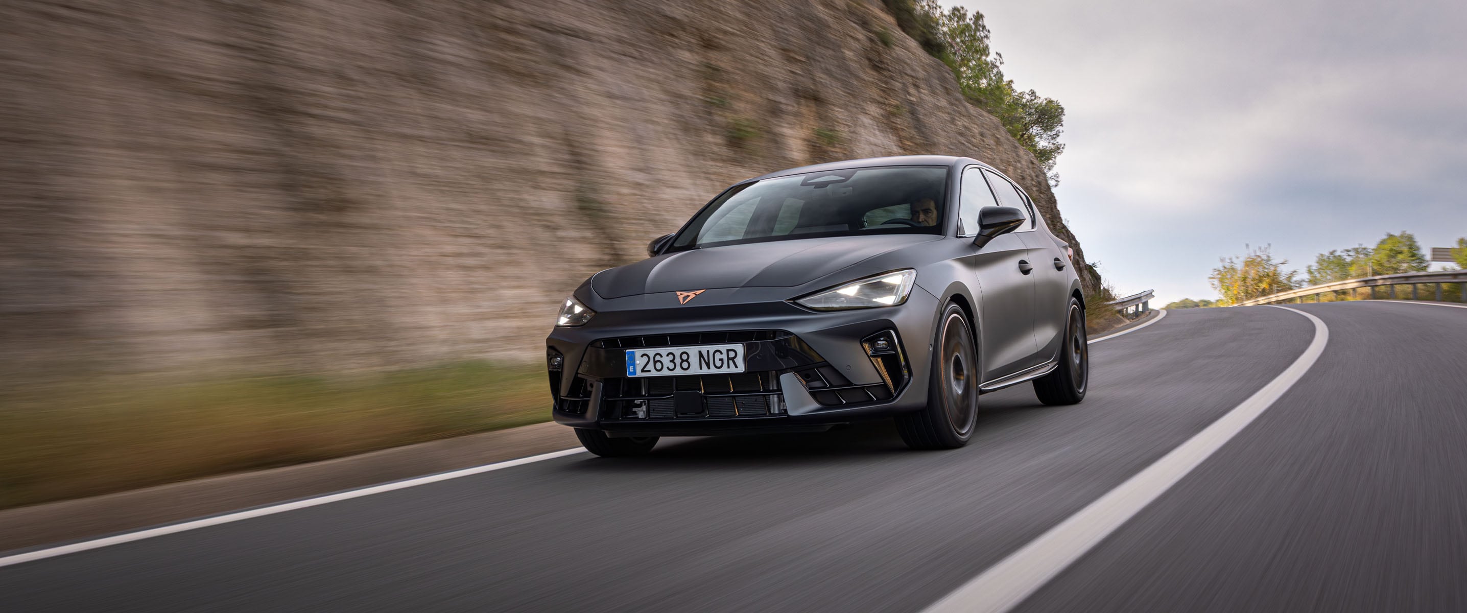 A CUPRA Leon VZ is driving quickly on a winding mountain road. The car is angled toward the camera, with motion blur indicating speed, and a rocky hillside cliff visible on the left.