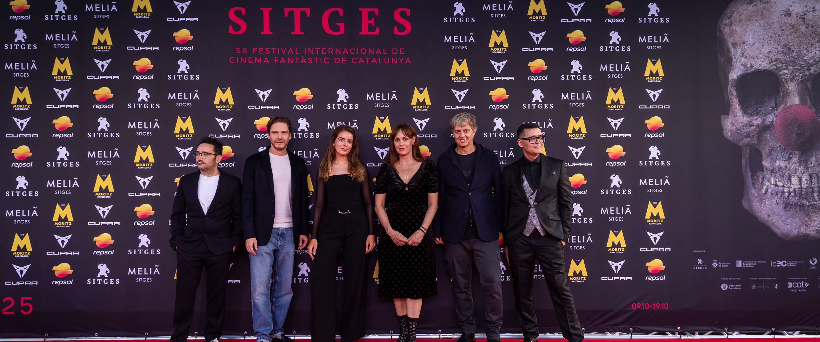 A group of six people, including actors and filmmakers, stand on the red carpet at the Sitges Film Festival 2025. A large Sitges logo is behind them, with a sponsor wall and a skull graphic on the right.