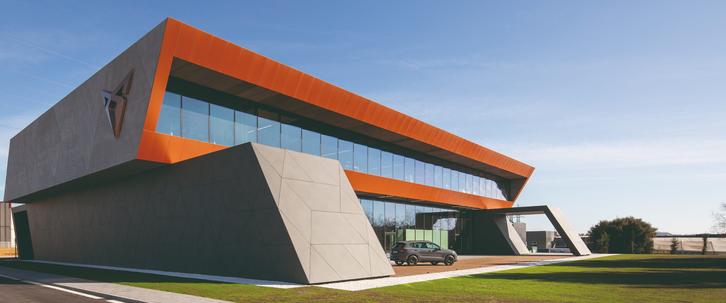 A modern, angular building with a prominent orange roofline and the CUPRA logo displayed on its facade, set against a clear blue sky
