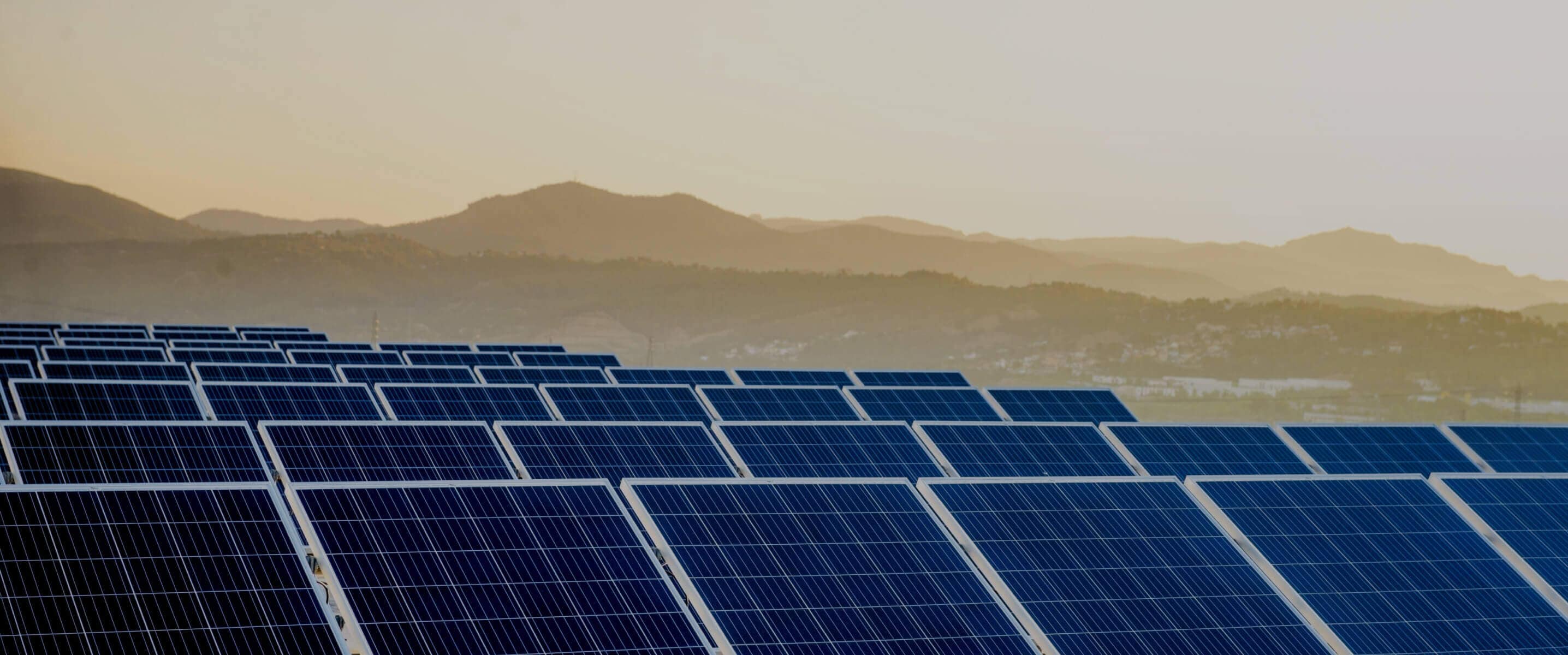 Rows of solar panels in the foreground with mountains and a hazy sunset in the background.