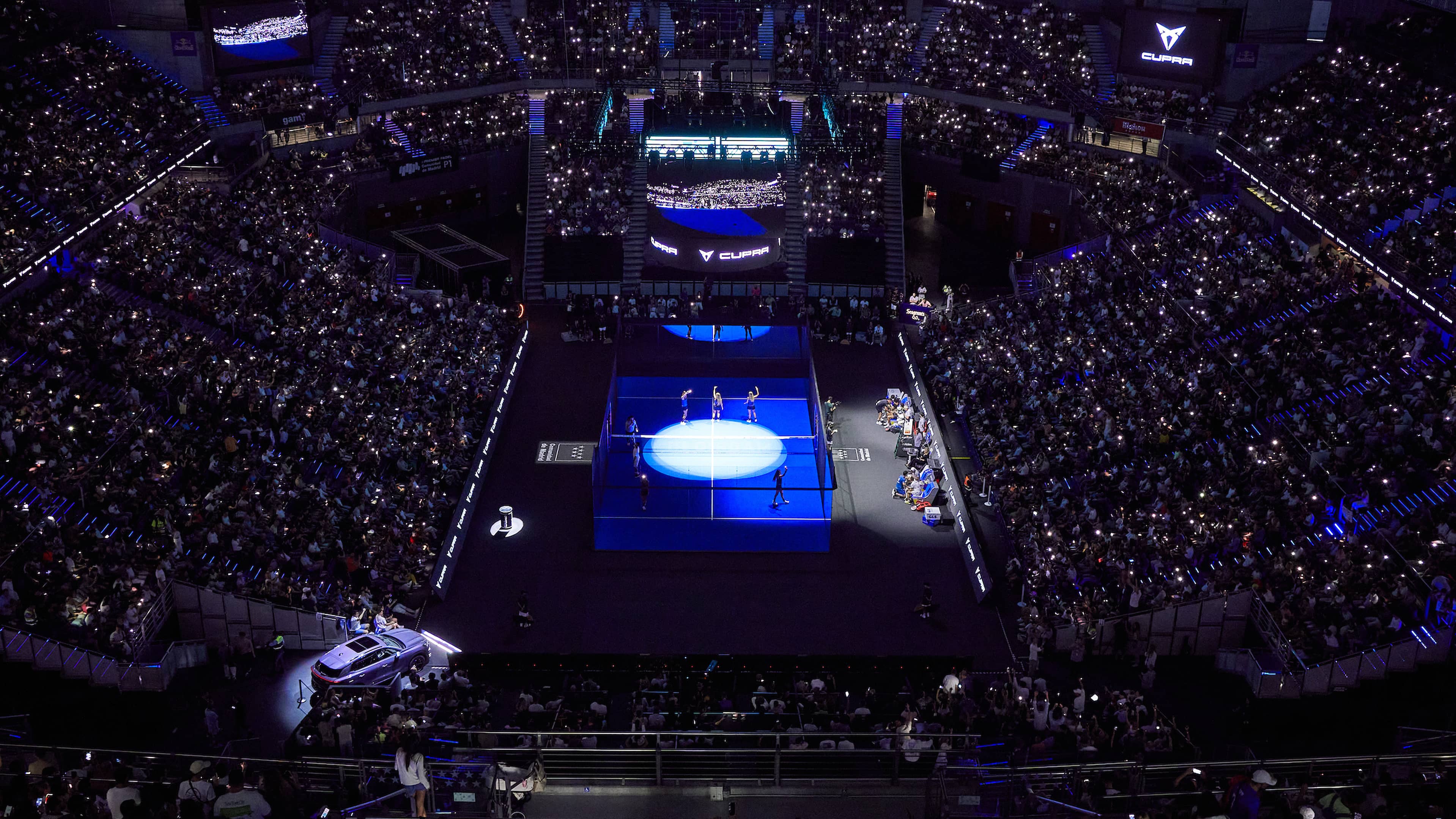 A large indoor stadium filled with spectators watching a padel match on a brightly lit court surrounded by glass walls. The audience is seated in tiers, and the court is the main focus under strong lighting.