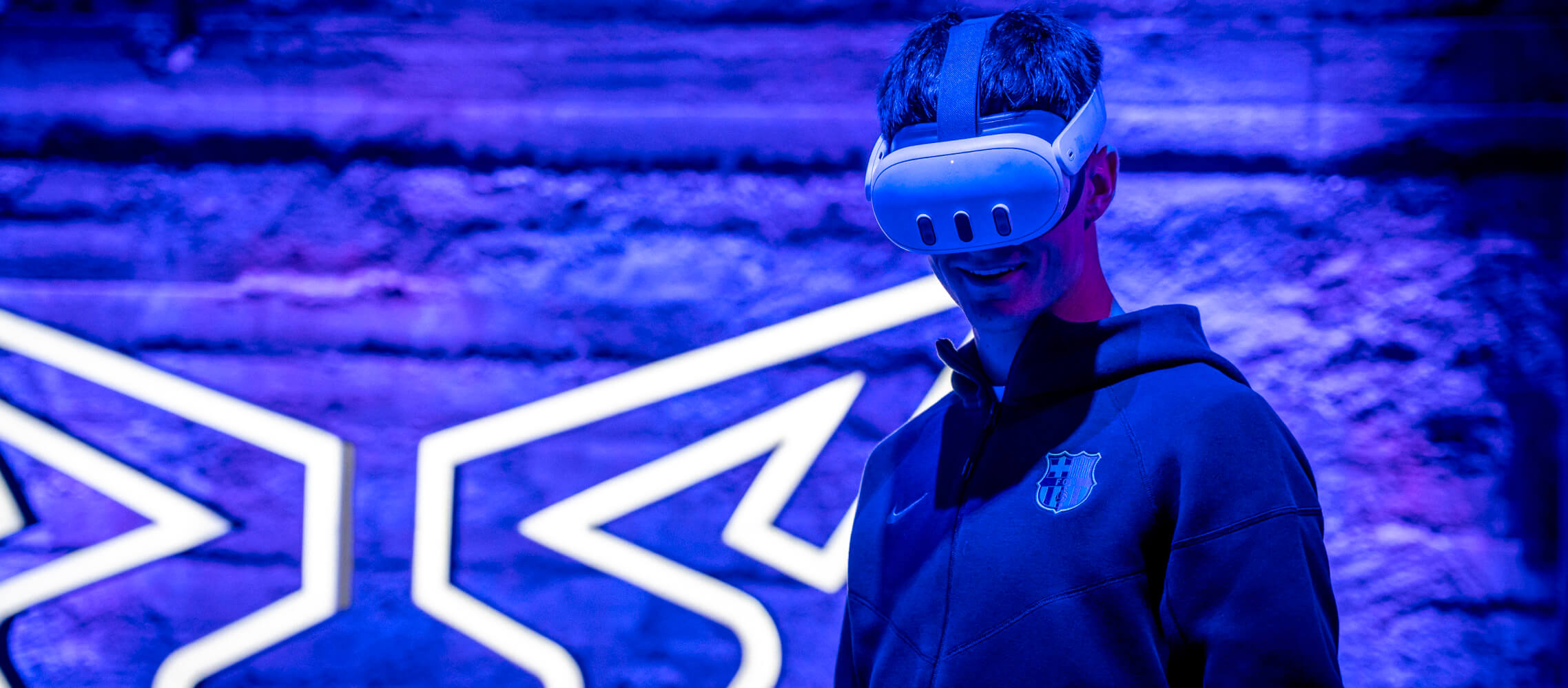 A FC Barcelona player wearing a virtual reality headset stands in front of a neon-lit background featuring abstract geometric designs.
