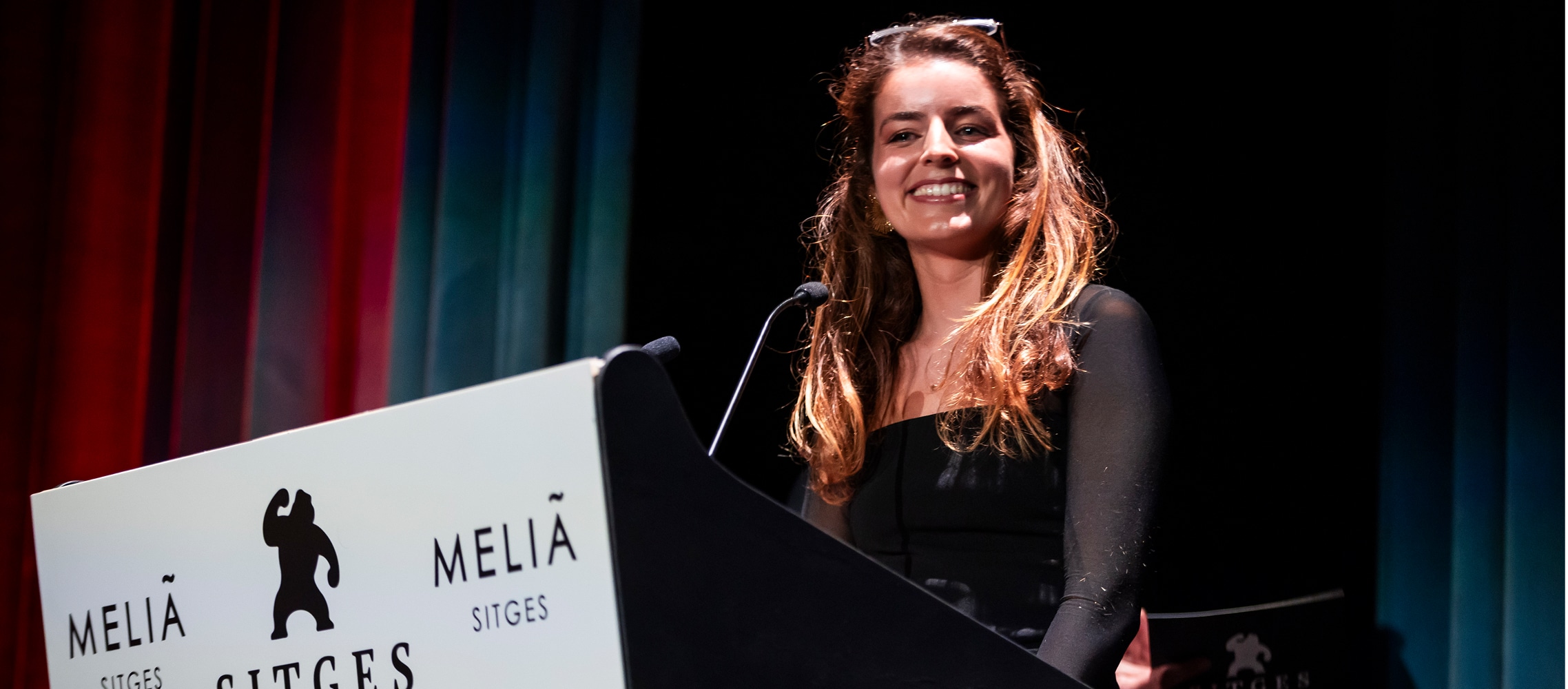 Martha Ayerbe is smiling as she stands behind a white podium with the black SITGES Film Festival and Meliá logos. She is speaking into a microphone on a stage with dark curtains in the background.