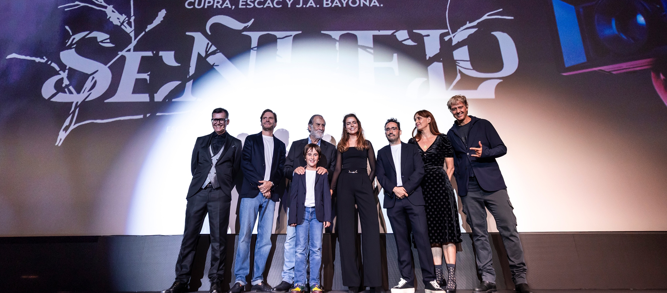A group of seven adults and one child stand on a stage in front of a large screen. The screen displays the title "The Dream Makers: A New Generation of Filmmakers" and credits the project as a collaboration between CUPRA, ESCAC, and J.A. Bayona.