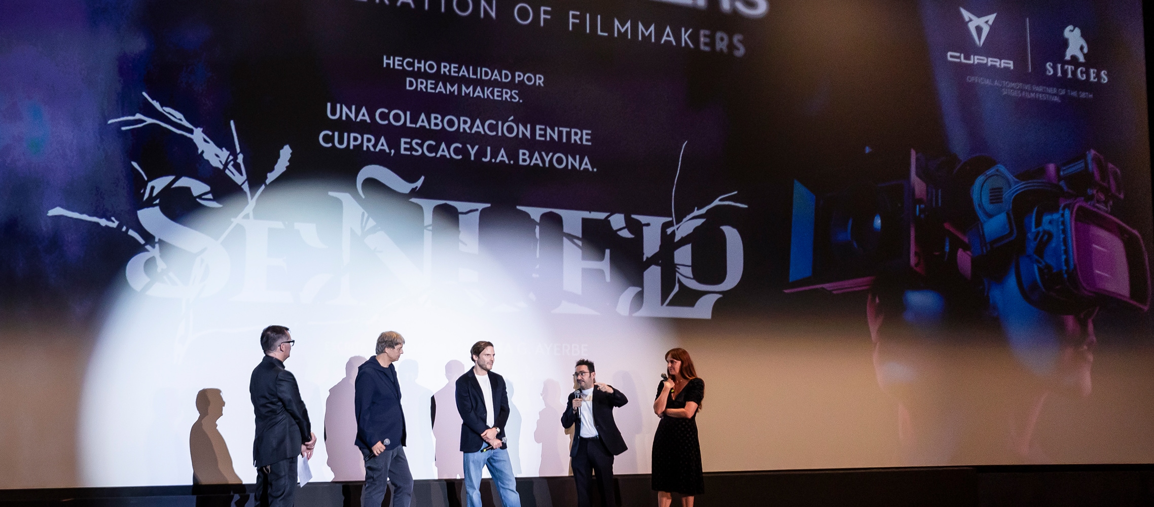 Five people are standing on a stage in a dark cinema, presenting "The Dream Makers: A New Generation of Filmmakers." The large screen behind them displays the title, the CUPRA and Sitges logos, and text indicating a collaboration between CUPRA, ESCAC, and J.A. Bayona.