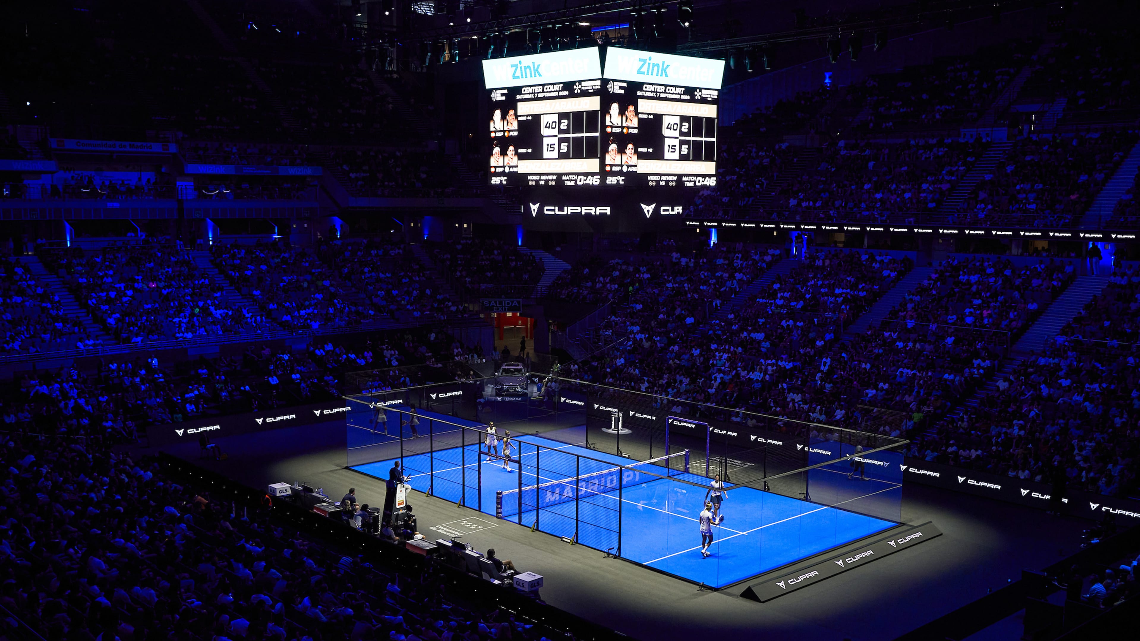 An overhead view of a brightly lit padel court during a match, surrounded by a large, dark stadium with spectators in the stands and a scoreboard visible above.