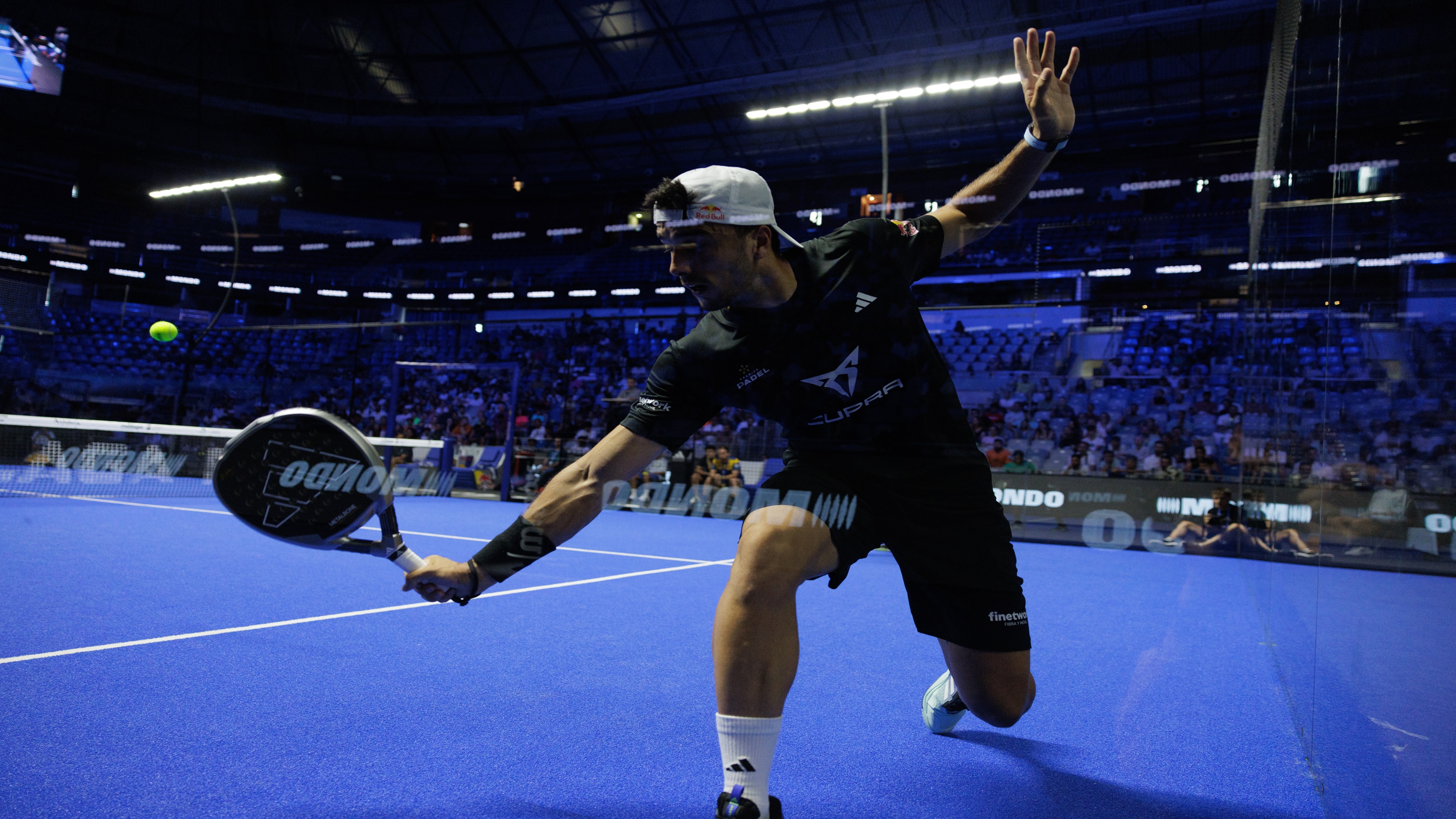 A padel player in action on a blue court, wearing dark sportswear and a white cap, mid-swing with a black padel racket.