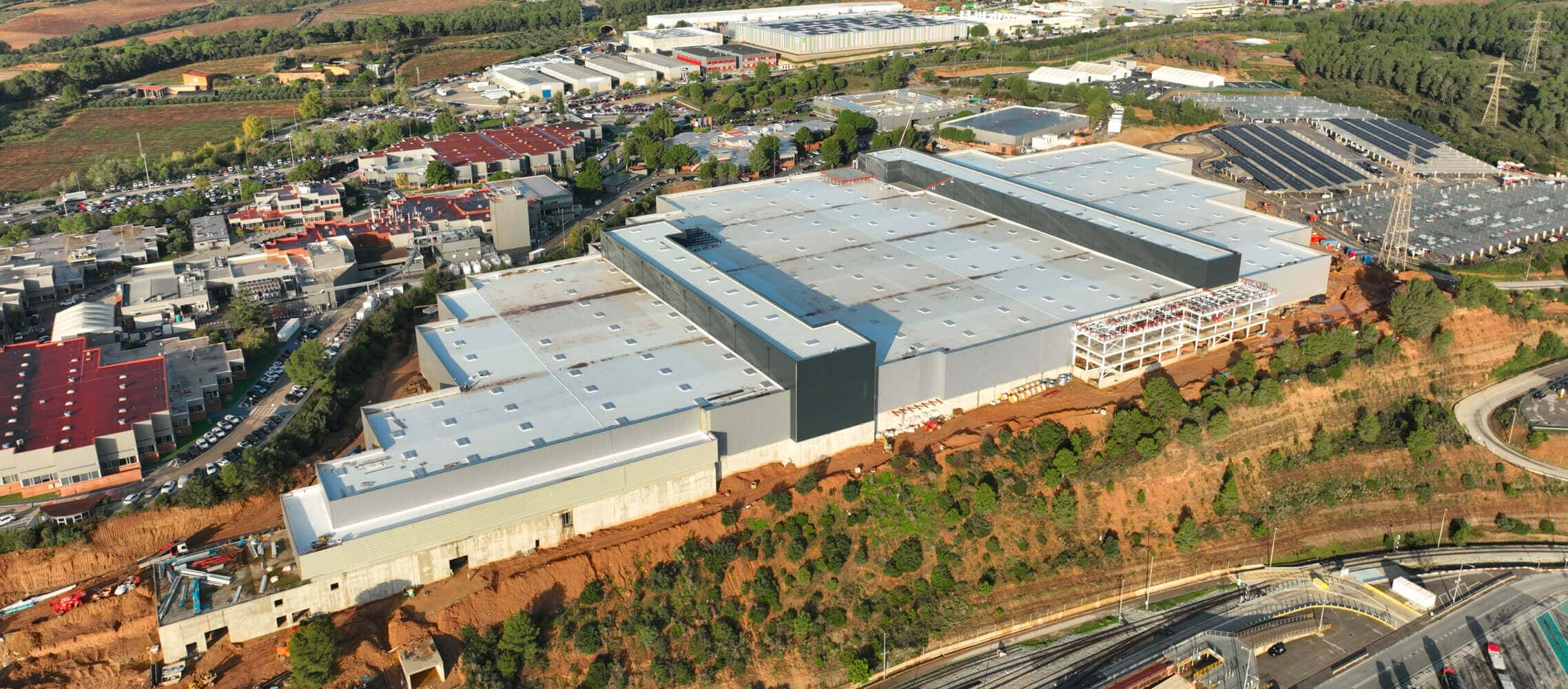 An aerial shot showcases a sprawling factory complex with multiple large buildings and extensive rooftops and roadways under a partly cloudy sky.