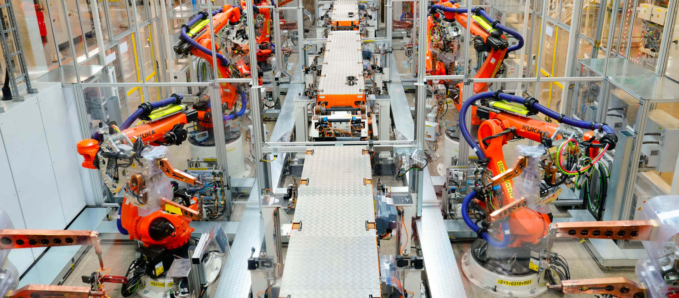 Overhead view of a modern car assembly line featuring multiple orange robotic arms working on vehicle chassis moving along a conveyor belt in a brightly lit factory setting.