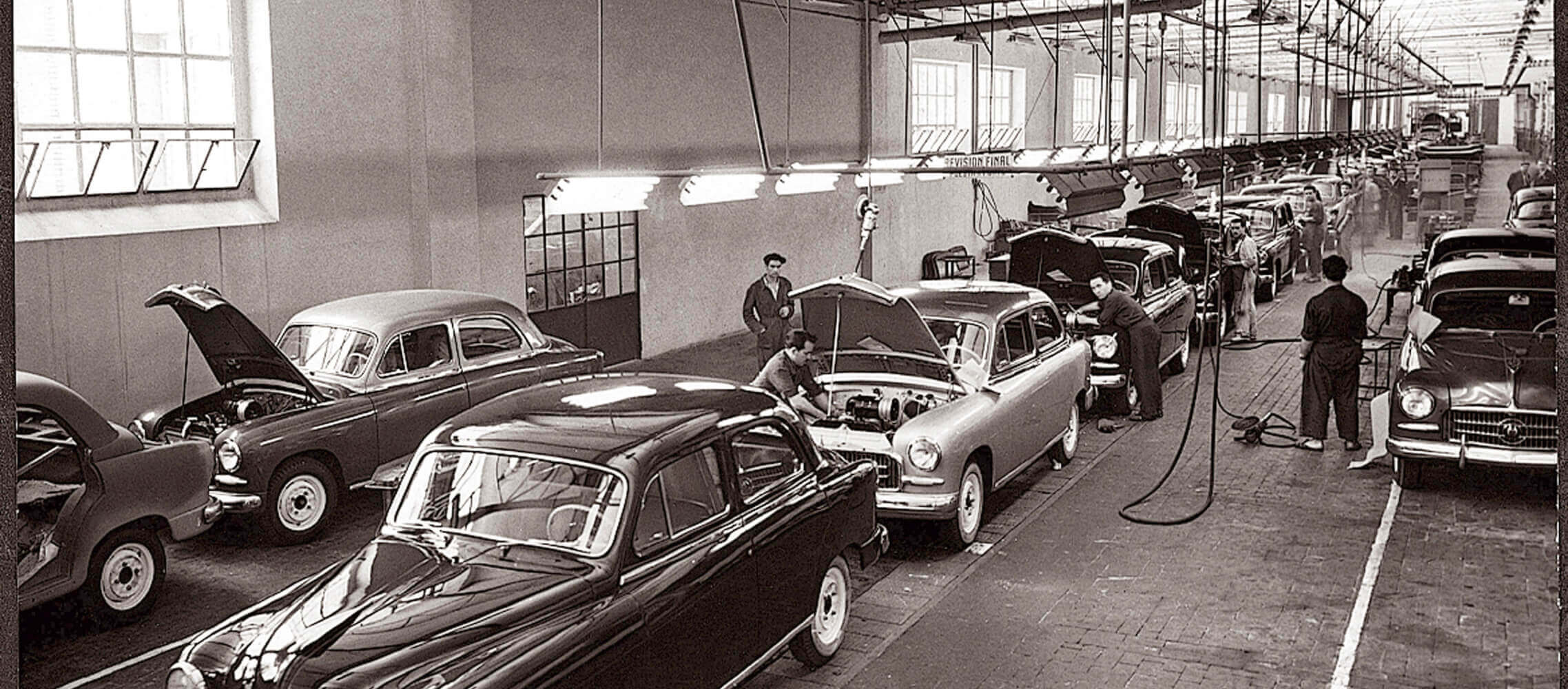 Vintage car assembly line with workers and classic cars in a factory.