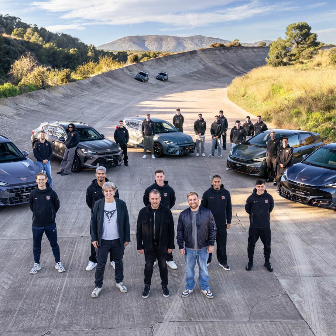 A football team and automotive brand representatives, pose together in front of several CUPRA vehicles on a sloped track surrounded by nature.