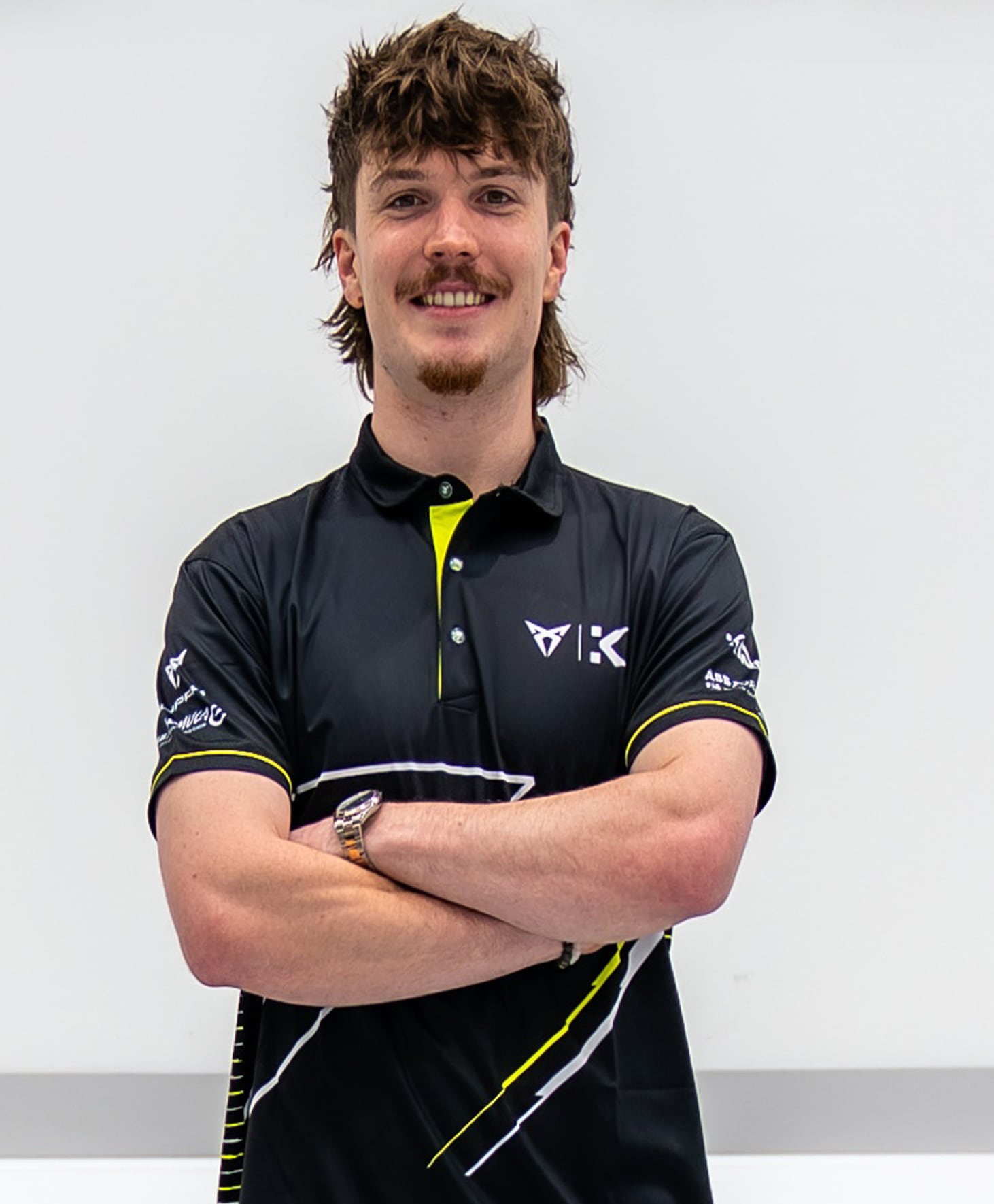 Professional racing driver Dan Ticktum stands with his arms crossed in front of a white wall with the black KIRO RACECO logo and the CUPRA logo, wearing a black and yellow team polo shirt.