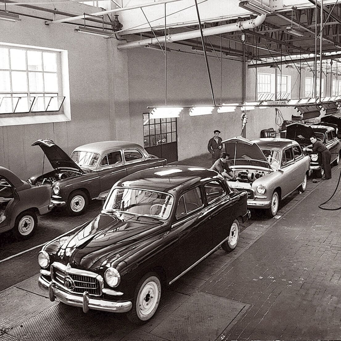 Vintage car assembly line with workers and classic cars in a factory.