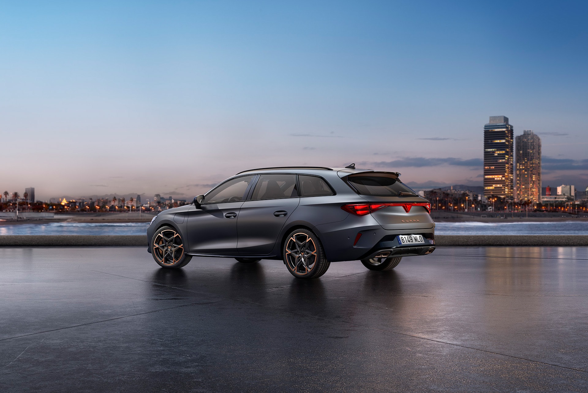 The sporty new 2025 Cupra Leon Sportstourer parked on concrete with a city skyline illuminated at twilight in the background. The vehicle is depicted in a matte grey finish.