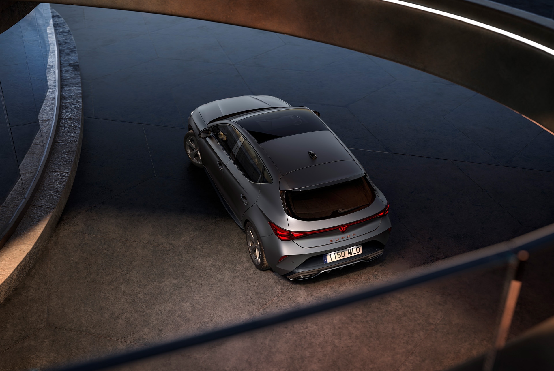 A top-down view of a sleek, matte grey CUPRA Leon. The car is parked on a dark surface. The car has a panoramic sunroof and distinctive alloy wheels. 