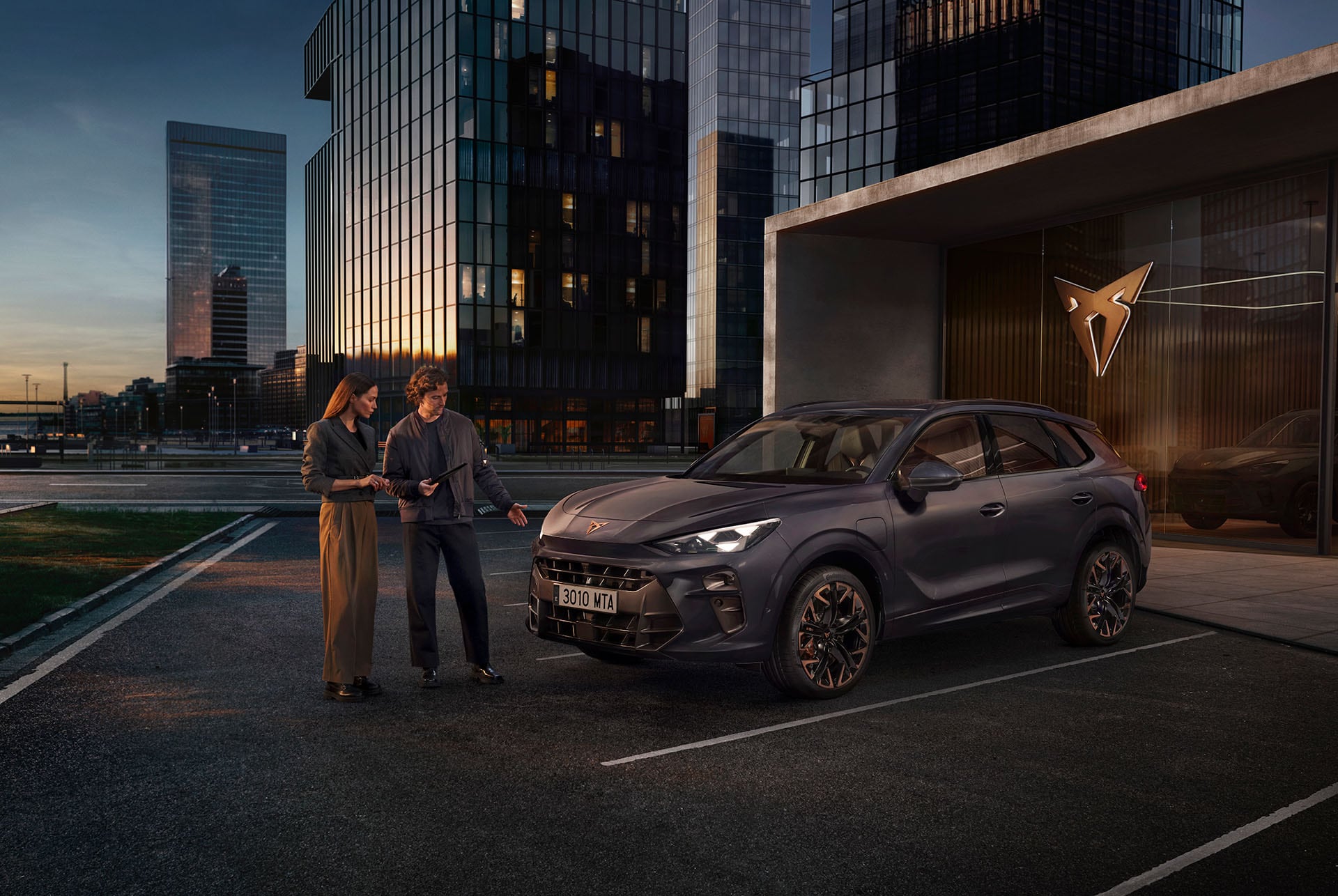 Two individuals standing in front of the CUPRA Terramar stationed in a parking next to a CUPRA building, in a city landscape. 