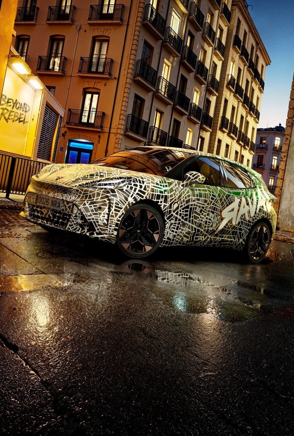 camouflaged CUPRA Raval with abstract white and green patterns parked on a wet urban street at dusk, surrounded by tall apartment buildings with lit windows.