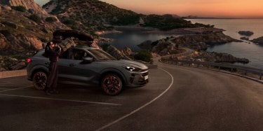 A CUPRA Terramar parked at a scenic mountain location with snow-covered peaks and winding roads in the background. A person stands next to the car, which is equipped with a roof box.