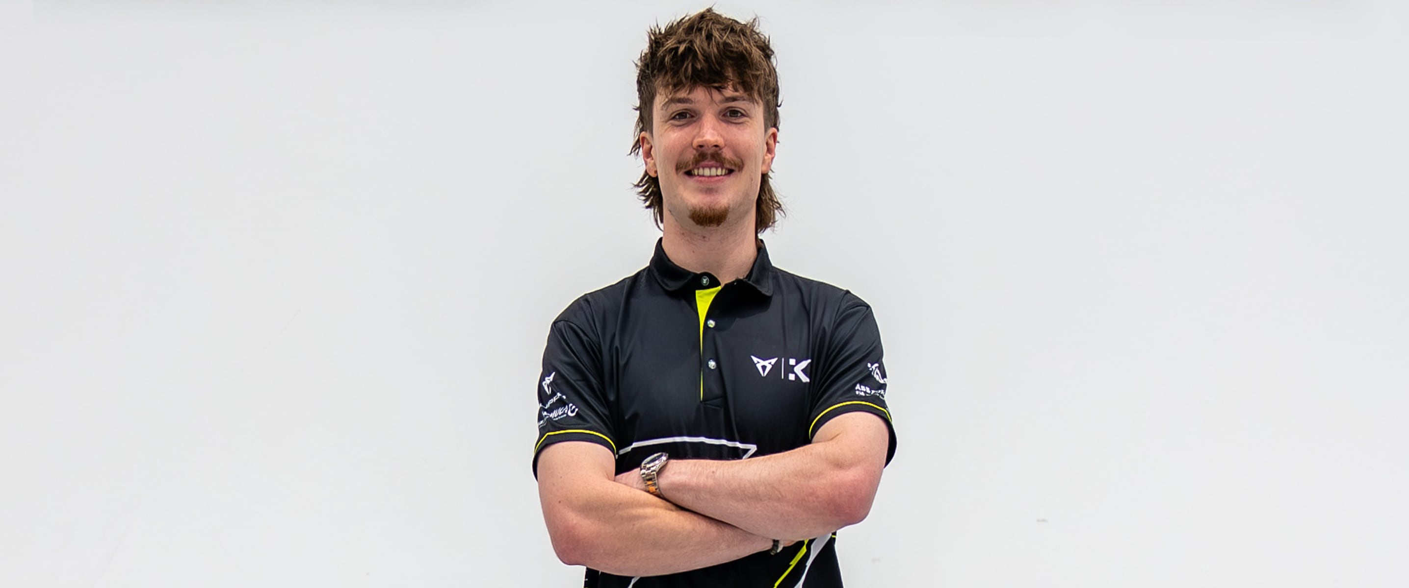 Professional racing driver Dan Ticktum stands with his arms crossed in front of a white wall with the black KIRO RACECO logo and the CUPRA logo, wearing a black and yellow team polo shirt.