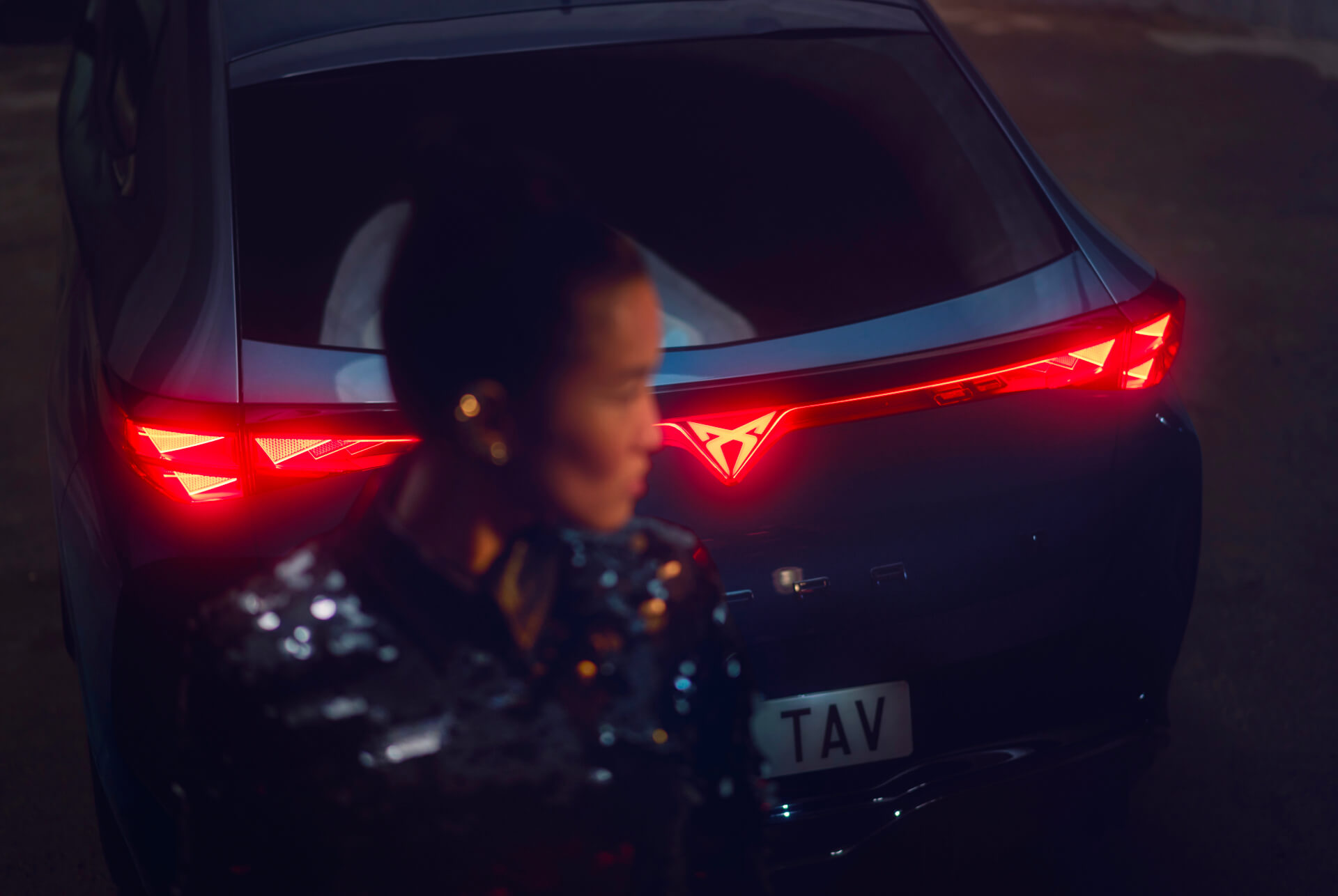 Rear view of a CUPRA vehicle featuring illuminated tail lights and the brand's distinctive glowing logo in the center; a woman in the foreground is slightly out of focus.