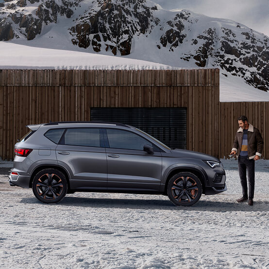 A man in a winter jacket looks down at his smartphone, standing beside the driver's door of a CUPRA Ateca. The car is parked on snow-dusted ground in front of a modern wooden building, with a vast, snow-covered mountain range in the background.