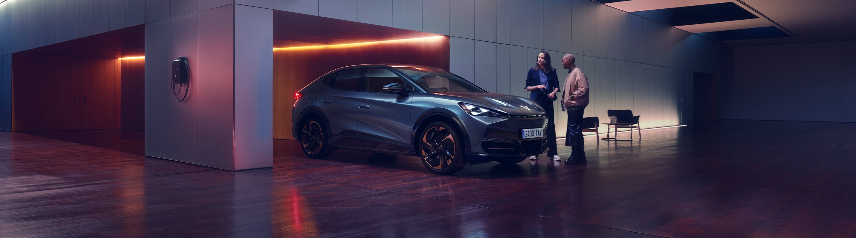 A blue CUPRA Tavascan in a front three-quarter view within a modern garage. Two people stand beside the vehicle and a wall charger, next to a copper CUPRA logo.