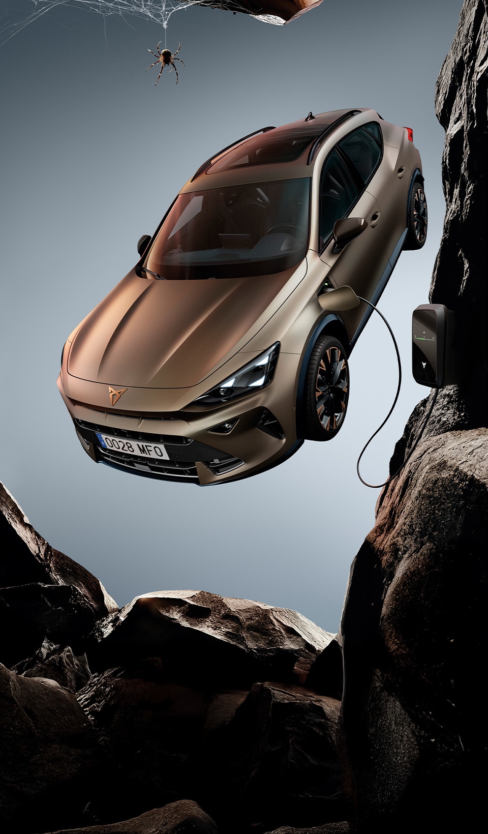 A CUPRA Formentor CUV suspended diagonally, next to a rocky, cliff-like structure. A charging cable is connected to the car from a charging unit on the cliff face.