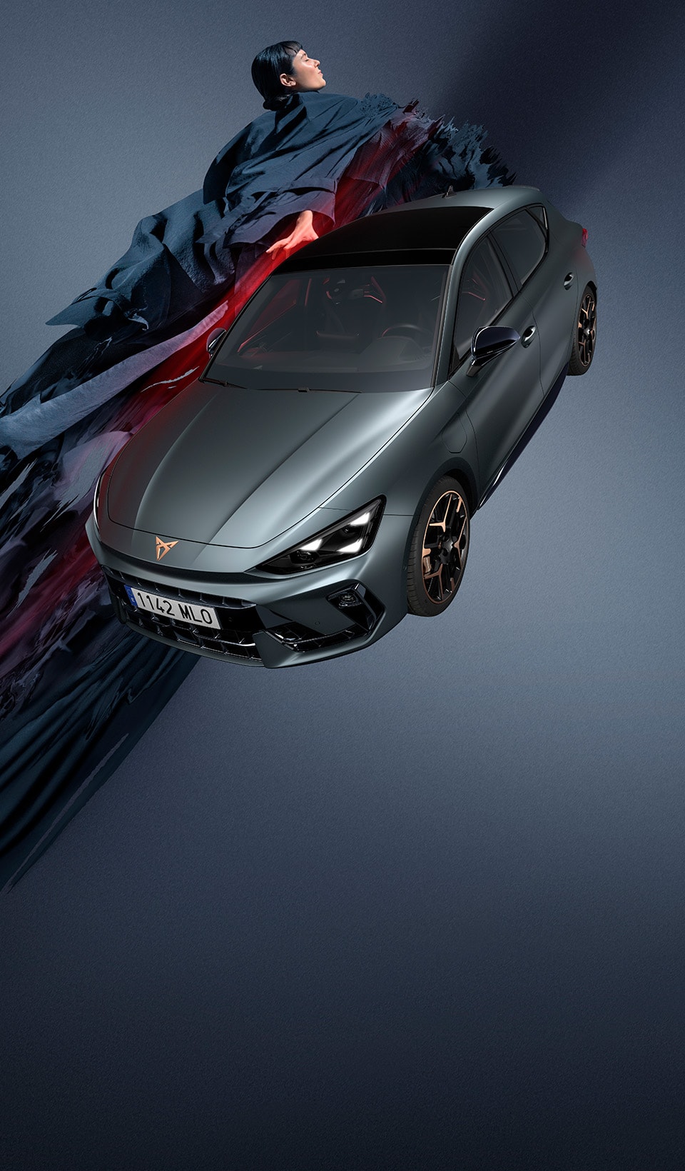 A CUPRA Leon hatchback seen from an elevated angle with its headlights on, next to a person covered in dark, flowing fabric that drapes onto the ground.