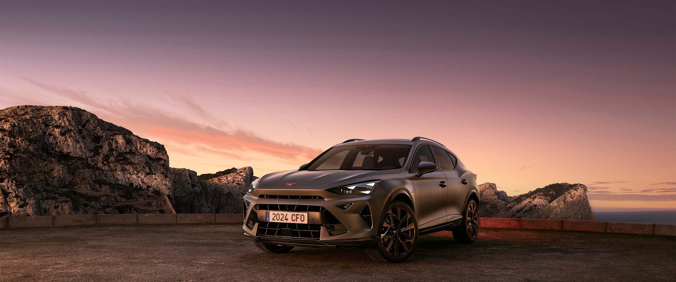 A three-quarter view of the new CUPRA Formentor 2024 parked on gravel at dusk, featuring a sleek, metallic body against a backdrop of rugged cliffs and ethereal sunset.