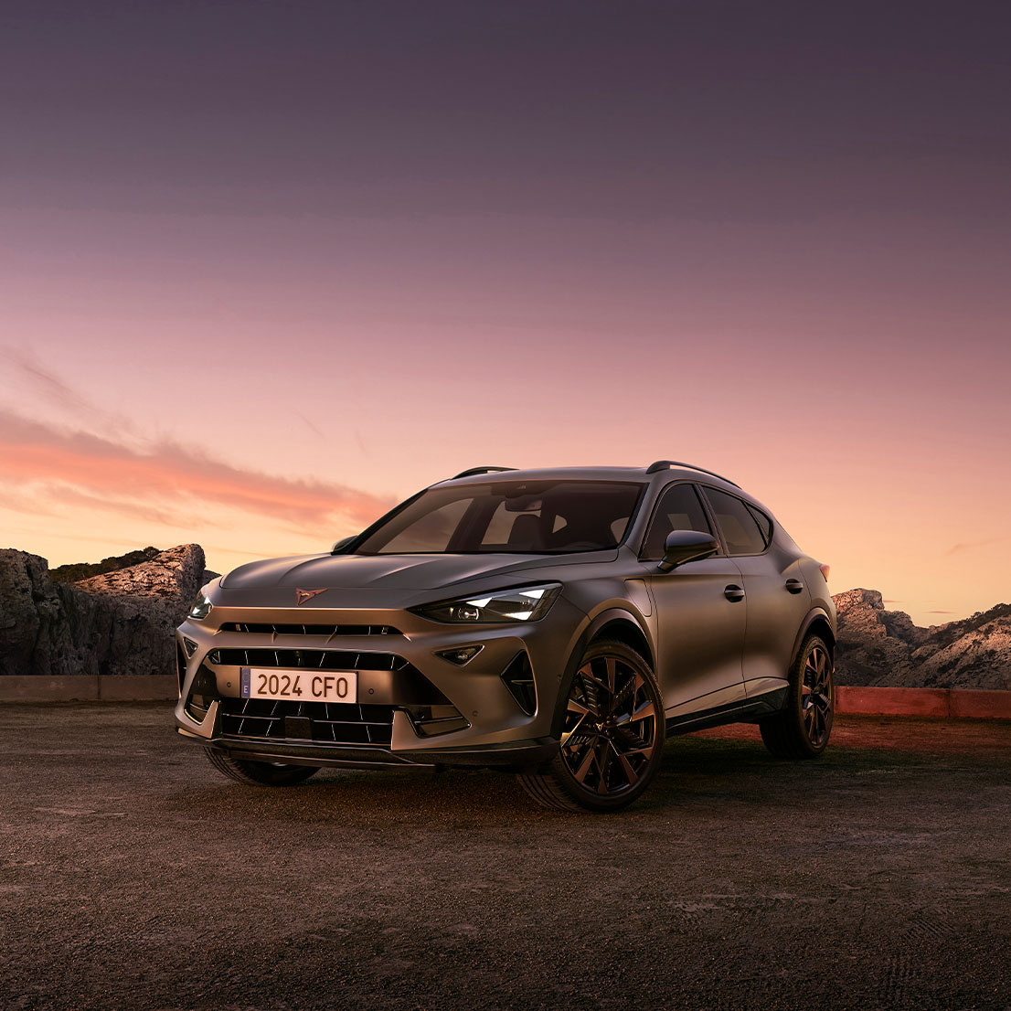 A three-quarter view of the new CUPRA Formentor 2024 parked on gravel at dusk, featuring a sleek, metallic body against a backdrop of rugged cliffs and ethereal sunset