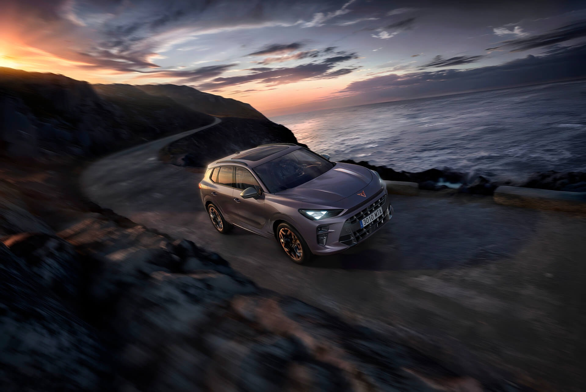 A dark gray Cupra Terramar SUV, front view, is parked on a reflective surface near a rocky shoreline at dusk or dawn. 